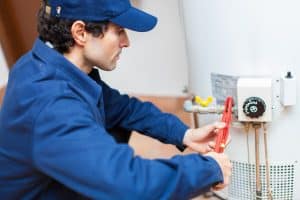 A plumber in a blue uniform uses a red pipe wrench to repair a wall-mounted water heater, inspecting the pipes and temperature control connections.